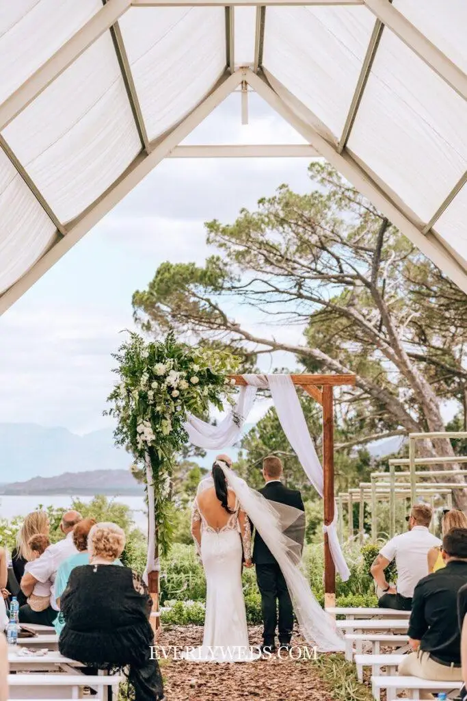 Outdoor wedding ceremony under wooden arbor with white draping and greenery - budget-friendly backyard wedding decor ideas