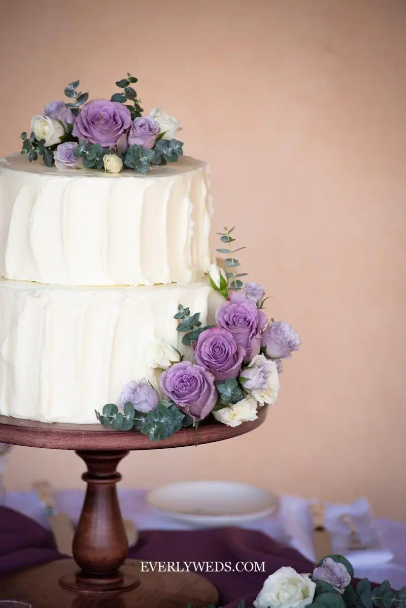 Simple two tier wedding cake classy design with smooth white buttercream and fresh flowers on a rustic wood slice at an outdoor wedding