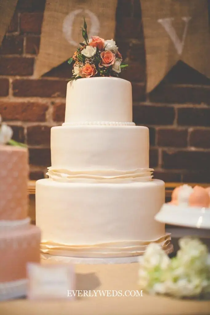 Simple three tier wedding cake elegant design with smooth buttercream and single flower cluster topper on a tall cake stand at a wedding reception