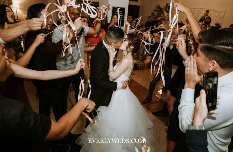 Bride and groom kissing surrounded by guests waving ribbon wands at wedding celebration