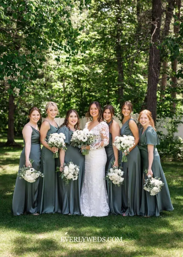 Bridesmaids wearing sage green bridesmaid dresses in different silhouettes holding white and blush floral bouquets at an outdoor green wedding theme ceremony